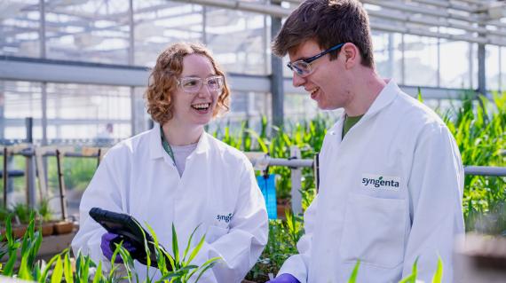 A man and woman in lab coats standing over some plants in a greenhouse.